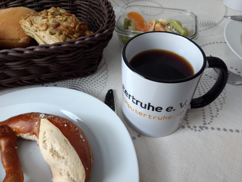 Eine mit Kaffee gefüllte Tasse mit dem Computertruhe-Schriftzug steht auf einem Frühstückstisch mit Gebäck und Obststückchen.