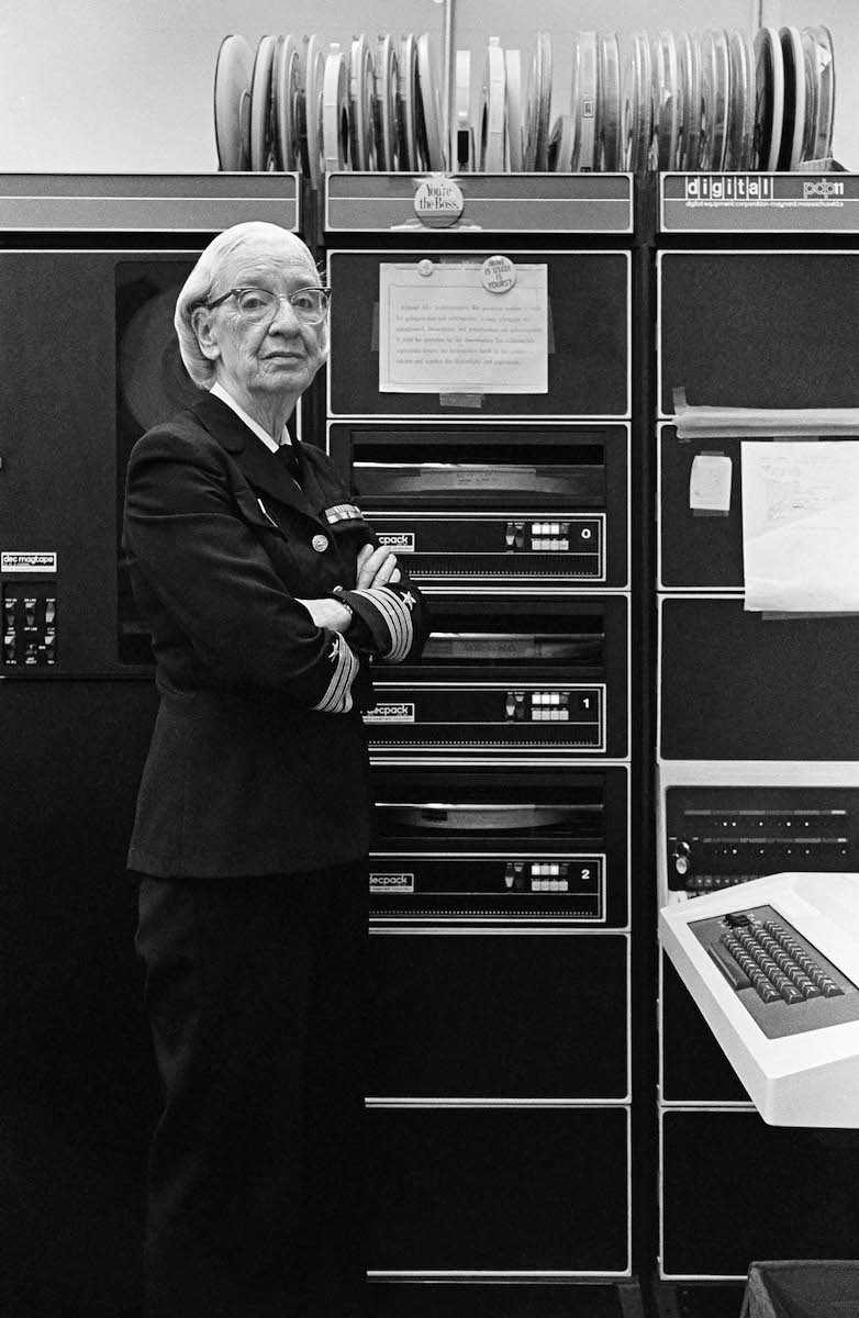 Grace Hopper in ihrer Navy-Uniform in ihrem Büro in Washington, D.C. Im Hintergrund sieht man eine PDP-11, im Vordergrund eine Tastatur.