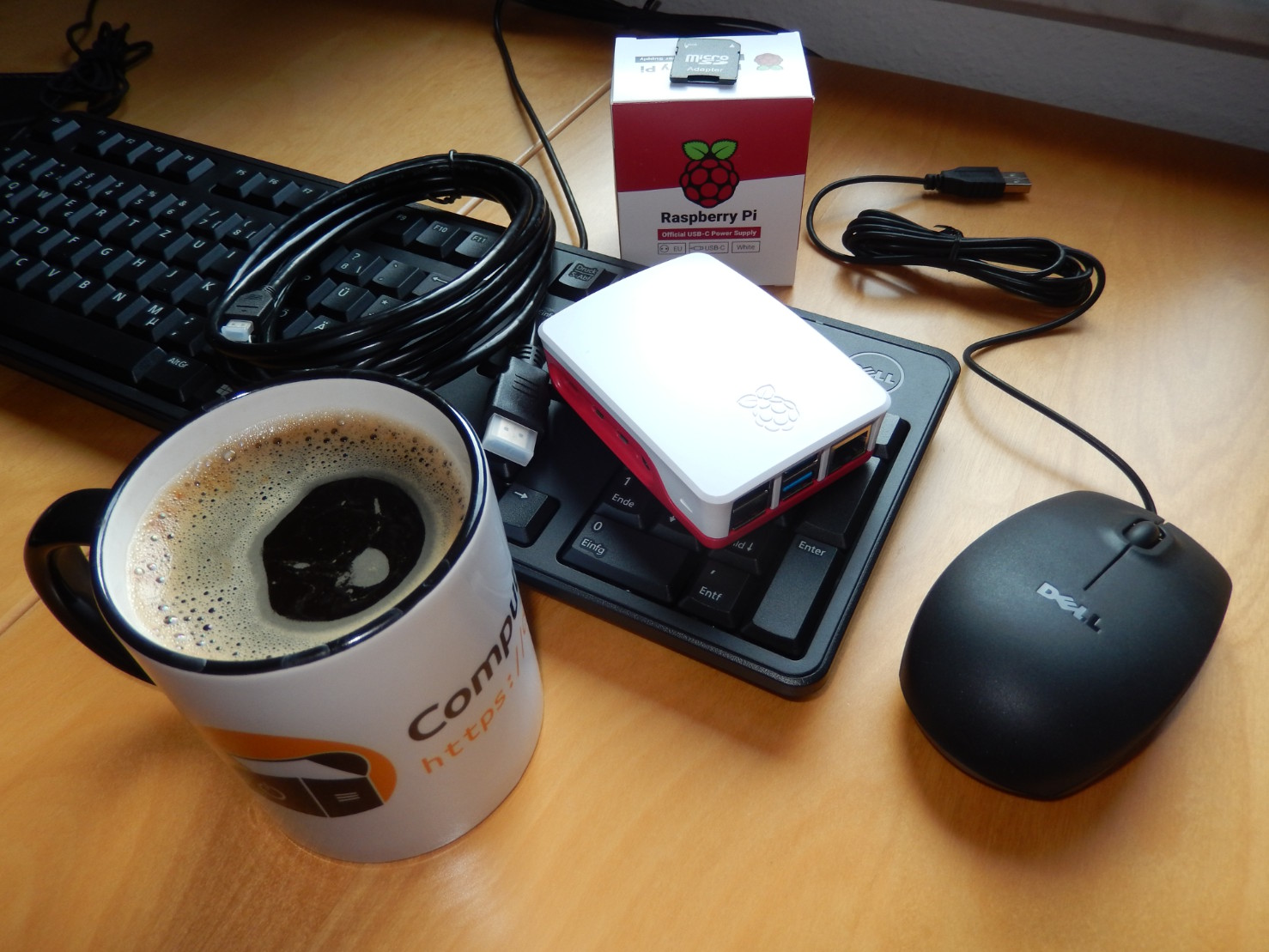 Eine Tasse mit dem „Computertruhe“-Logo und -Schriftzug, die mit Kaffee gefüllt ist. Zusätzlich liegen auf dem Tisch eine Tastatur, Maus, ein HDMI-Kabel, ein verpacktes Netzteil und ein „Raspberry Pi“ in einem Gehäuse.