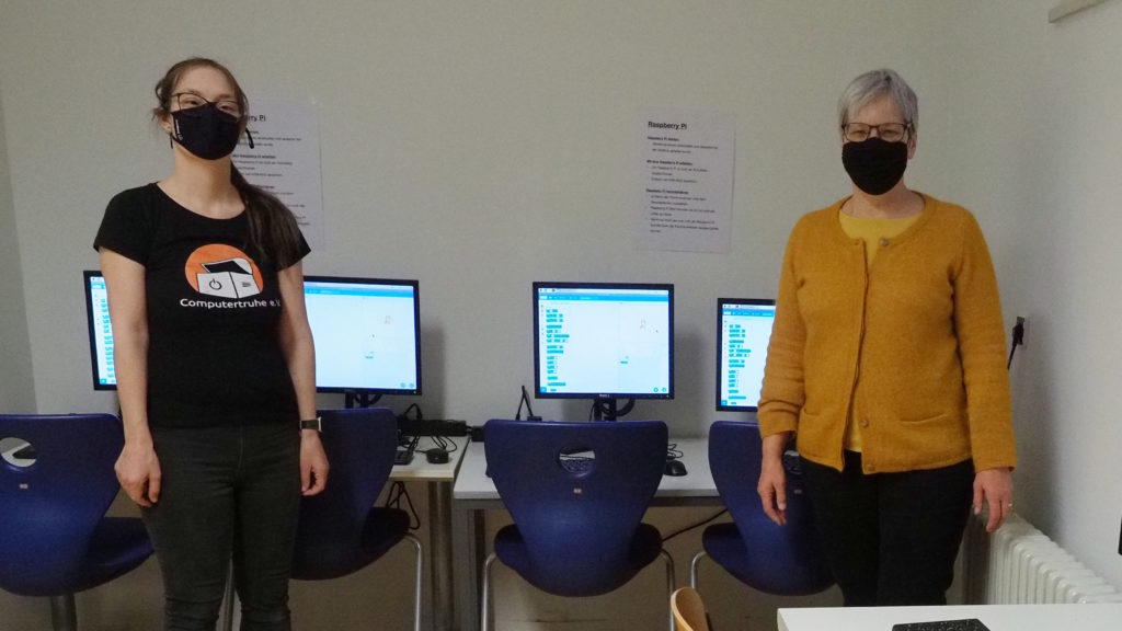 Julia Fiedler und Heidrun Albert stehen mit Abstand und Mund-Nasen-Schutzmasken im neuen Computertraum. Hinter ihnen befinden sich auf zwei Tischen verteilt vier Computerarbeitsplätze. Auf den eingeschalteten Monitoren lassen sich „Scratch“-Programme erahnen.