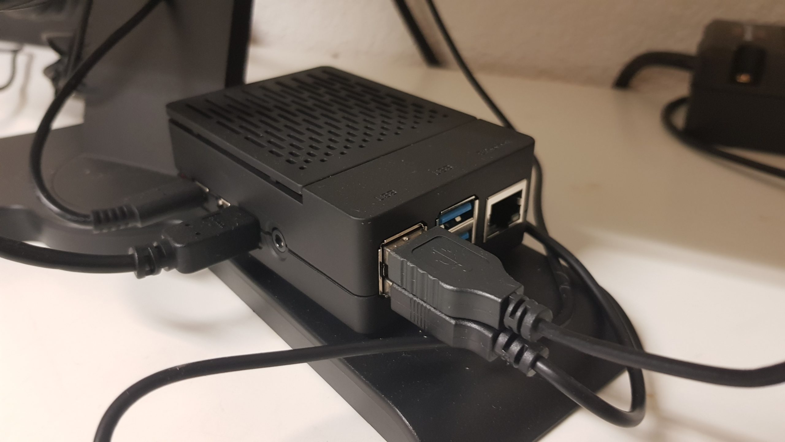Ein am Monitorstandfuss befestigter „Raspberry Pi“, der mit vier Kabeln verbunden ist. Er steckt in einem Gehäuse, bei dem der obere Teil abgenommen werden kann.