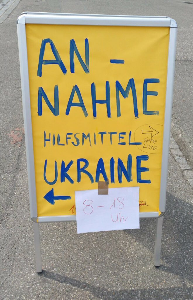 Ein Kundenstopper mit einem gelben Plakat, auf dem in blau geschrieben steht: „Annahme Hilfsmittel Ukraine (siehe Liste)“. Am unteren Ende klebt eine weiße DIN-A4-Seite mit dem Inhalt „8-18 Uhr“.