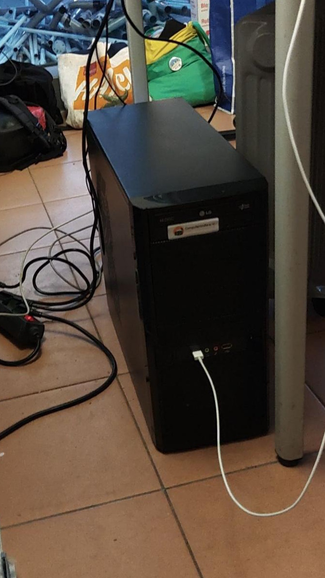 Auf einem gefliesten Boden steht neben einem Tisch ein schwarzer Desktop-Tower-PC, auf dessen Front ein Aufkleber der „Computertruhe“ klebt.