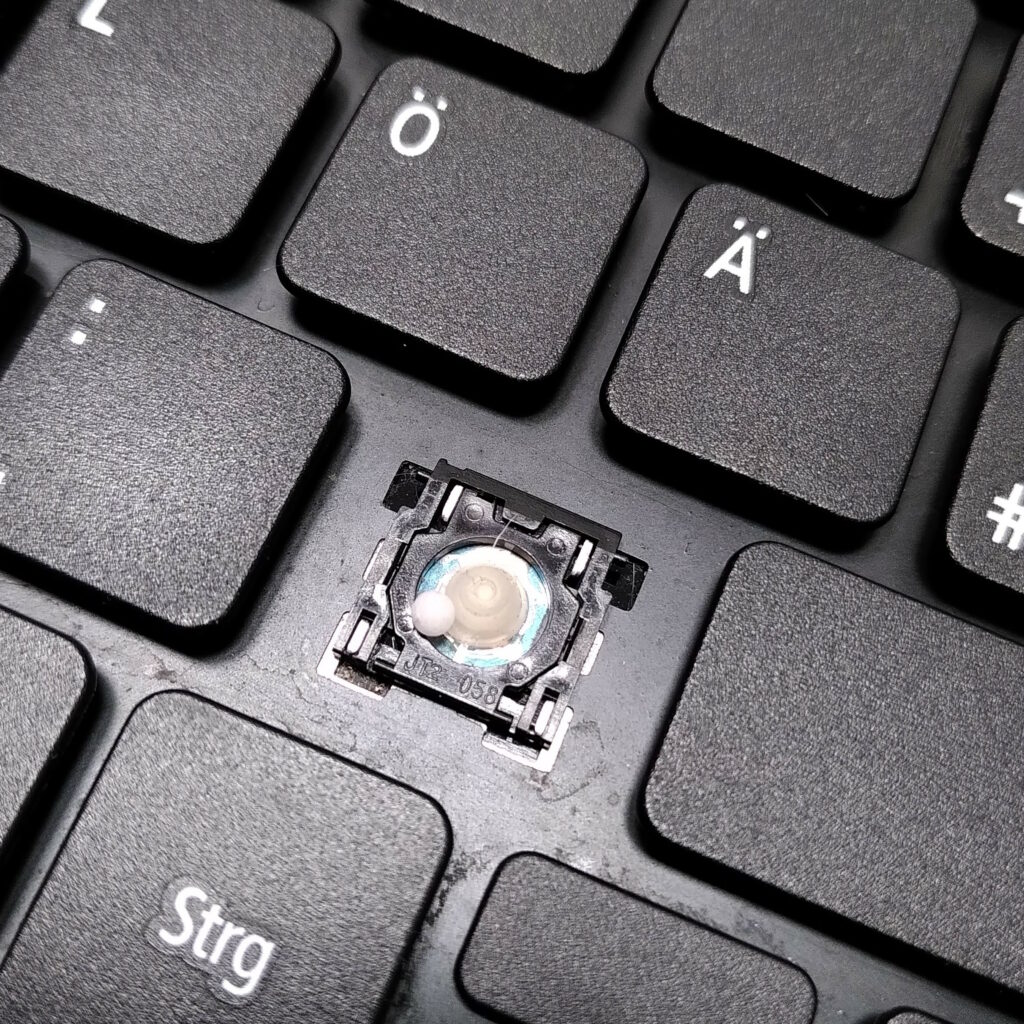 Eine Laptop-Tastatur, bei der die Tastenkappe der Minustaste fehlt. Neben dem Rubber-Dome liegt ein Zuckerglobulus.