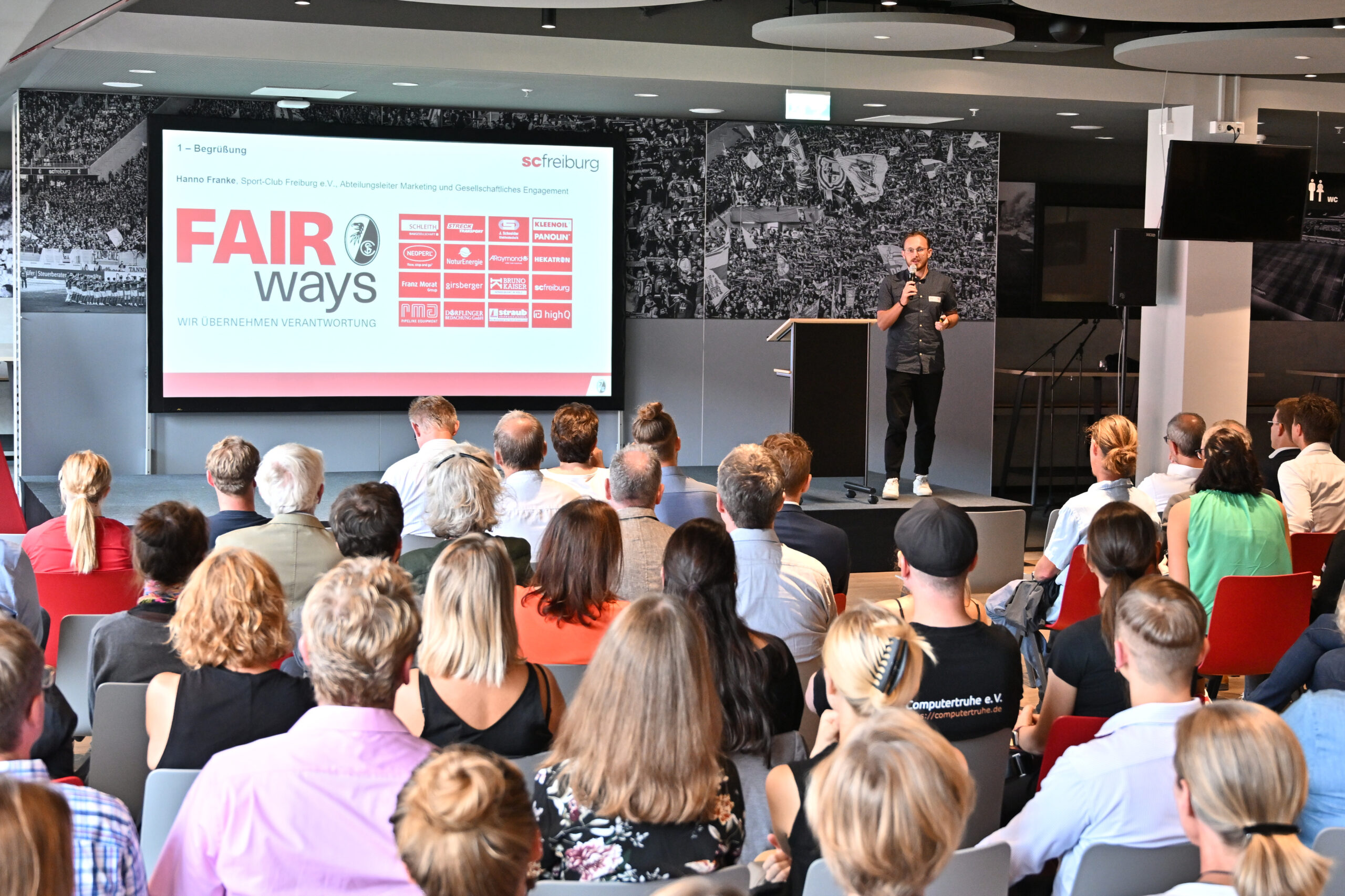 Tobias Rauber steht mit einem Mikrofon auf der Bühne. Neben ihm werden auf einer Leinwand die Logos der „FAIR ways“-Partner angezeigt. Im Vordergrund sieht man einen Teil des Publikums sitzen.