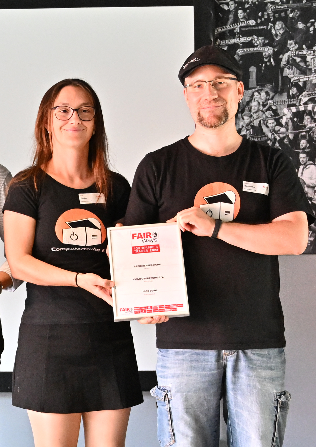 Julia Fiedler und Marco Rosenthal stehen lächelnd auf einer Bühne und halten gemeinsam eine gerahmte Urkunde in den Händen. Beide tragen ein schwarzes T-Shirt mit dem Logo der „Computertruhe“.