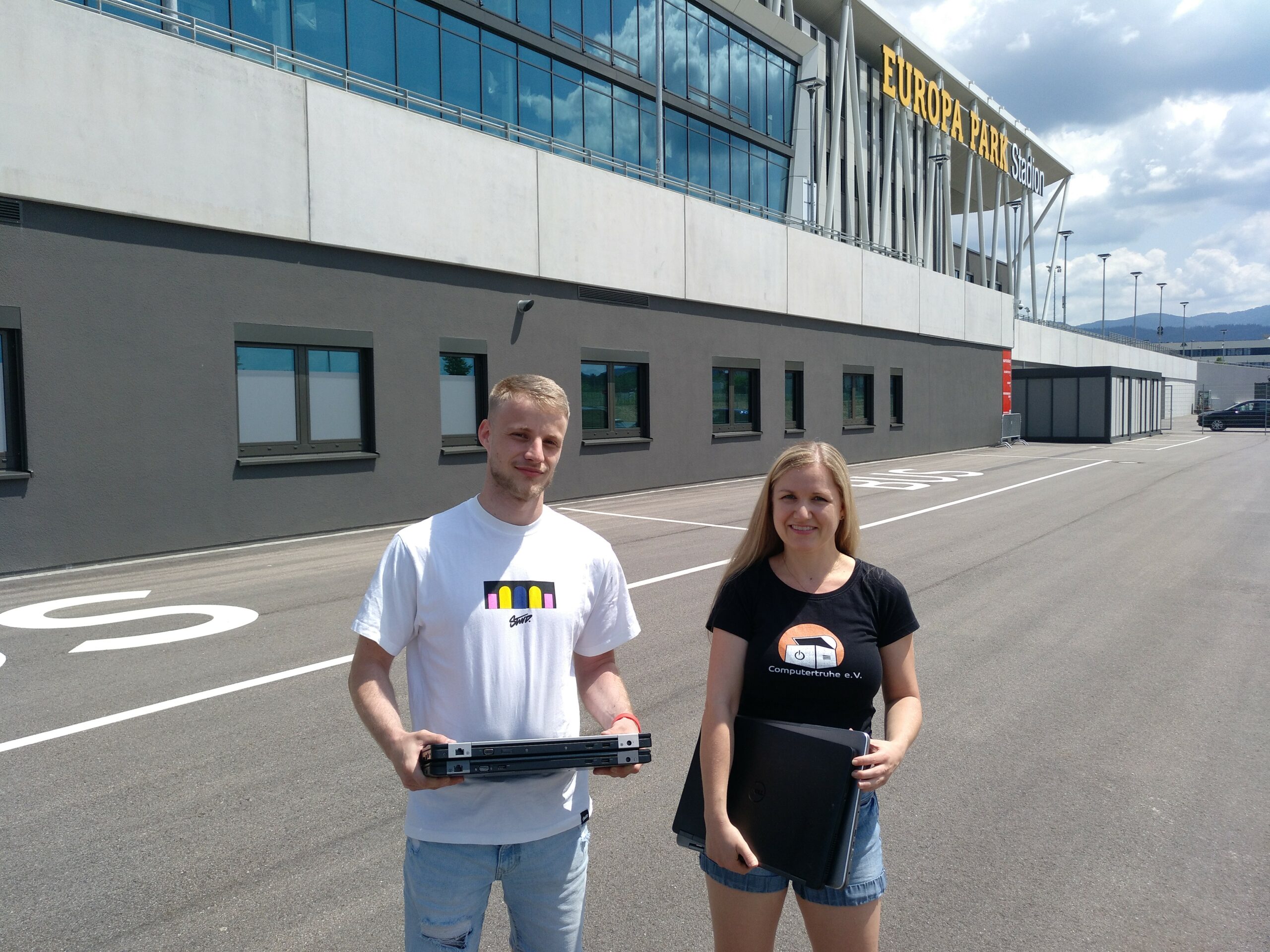 Auf dem Parkplatz, direkt vor dem Europa-Park Stadion, stehen Benedikt Auer und Annette Linder mit einigen Laptops in ihren Händen und lächeln in die Kamera.
