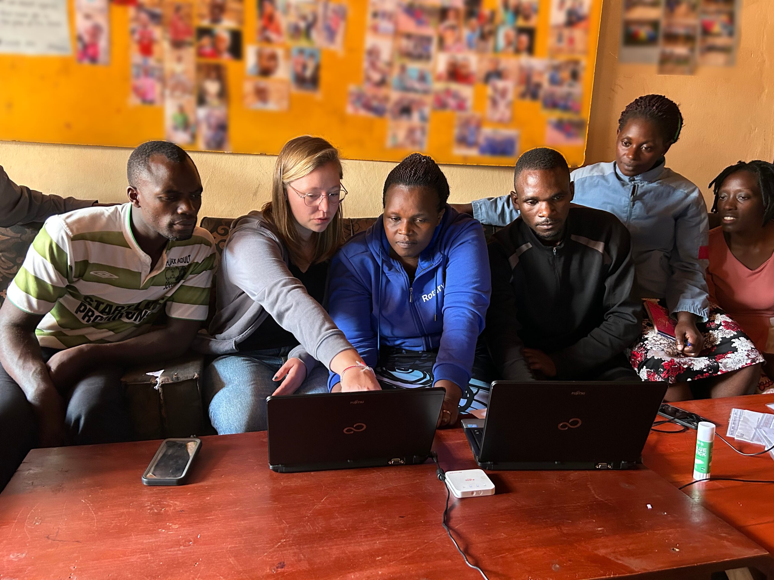 Sechs Personen sitzen vor einem Tisch, auf dem sich zwei aufgeklappte Laptops befinden. Eine hellhäutige Frau zeigt den anderen dunkelhäutigen Personen etwas auf einem der Bildschirme (Foto mit freundlicher Genehmigung des Vereins „KASA for Kids Uganda e. V.“).
