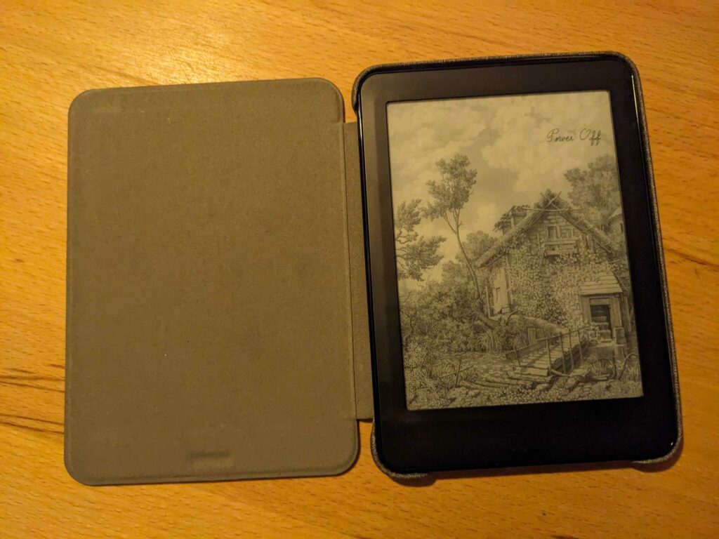 Foto des E-Book-Readers in einer aufgeklappten Schutzhülle. Das Display zeigt ein schwarz-weiß Bild eines kleinen Hauses mit Bach und Vegetation an.
