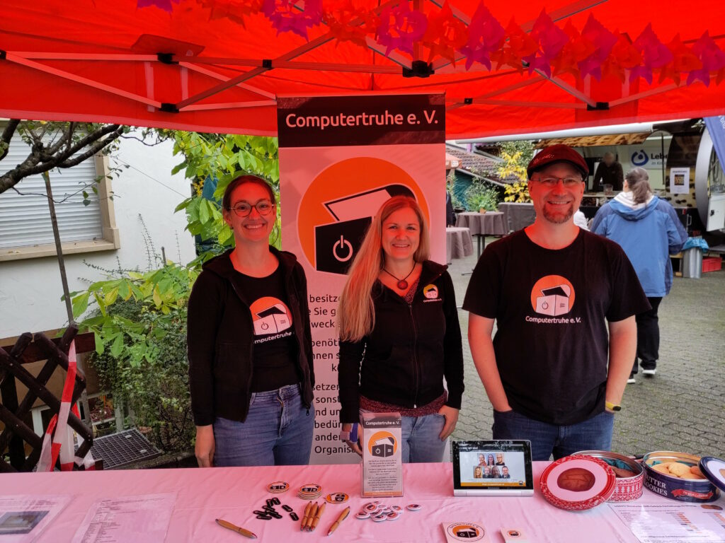 Foto zweier Frauen und eines Mannes unter einem orangefarbenem Pavillon. Die Personen lächeln und tragen schwarze Oberbekleidung, die das Vereinslogo ziert. Vor ihnen befindet sich ein langer Tisch mit diversem Info- und Werbematerial, einem Tablet, das eine Präsentation zeigt, und mit zwei geöffneten Keksdosen. Hinter ihnen steht ein Roll-up.