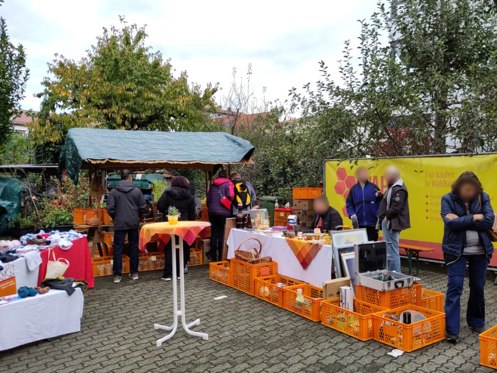 Foto das einige Verkaufsstände in einem gepflasterten Innenhof zeigt, die teils bedacht und teils unbedacht sind. Drumherum stehen Menschen, die die Stände betreuen oder die Waren begutachten.