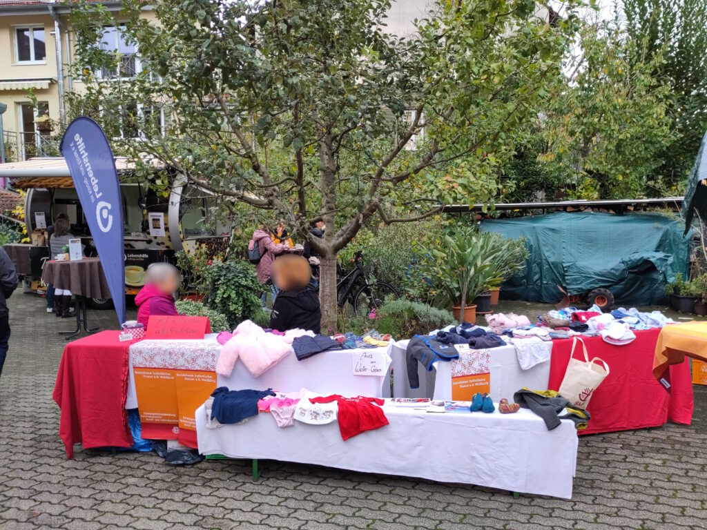 Foto des unbedachten und aus mehreren Tischen bestehenden Verkaufsstandes des Vereins „Sozialdienst katholischer Frauen e. V. Waldkirch“. Auf den Tischen befinden sich Kleidungsstücke für Kinder, dahinter sitzen zwei Personen.