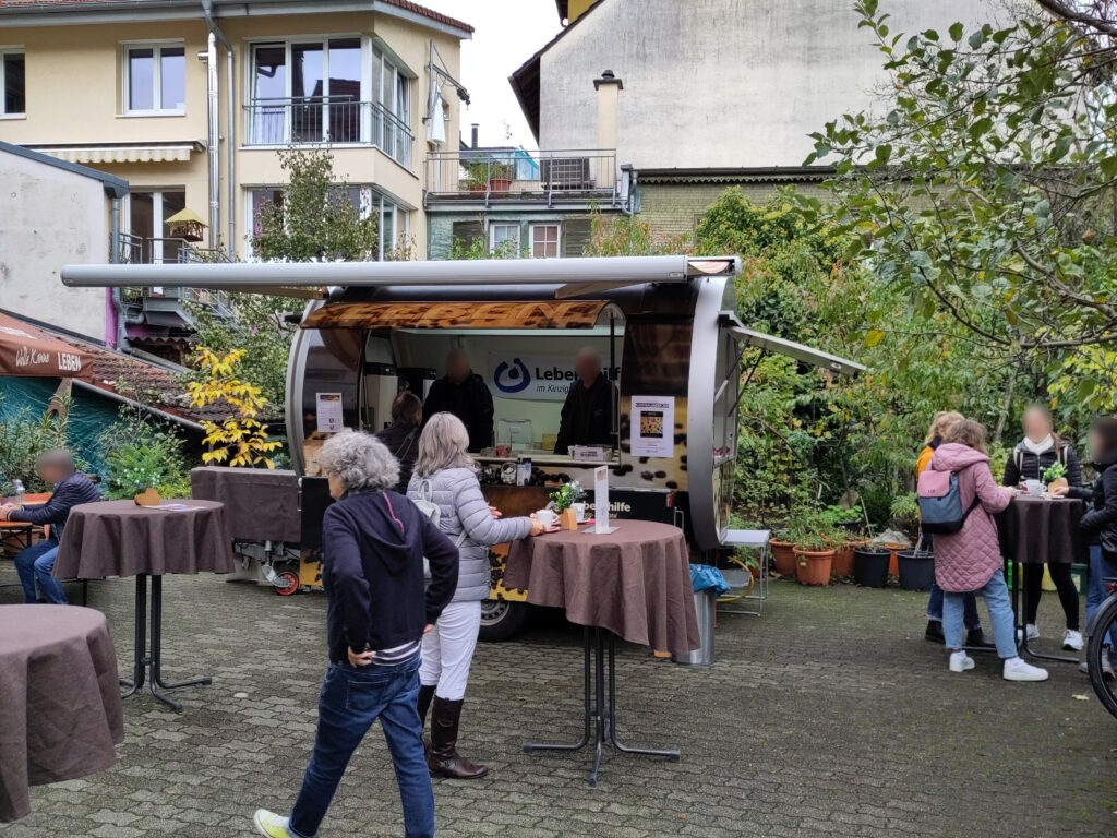 Foto eines Anhängers in Form einer Dose, in der zwei Personen stehen. Um den Hänger herum befinden sich Stehtische und weitere Menschen, die teilweise Kaffeetassen vor sich auf den Tischen stehen haben.