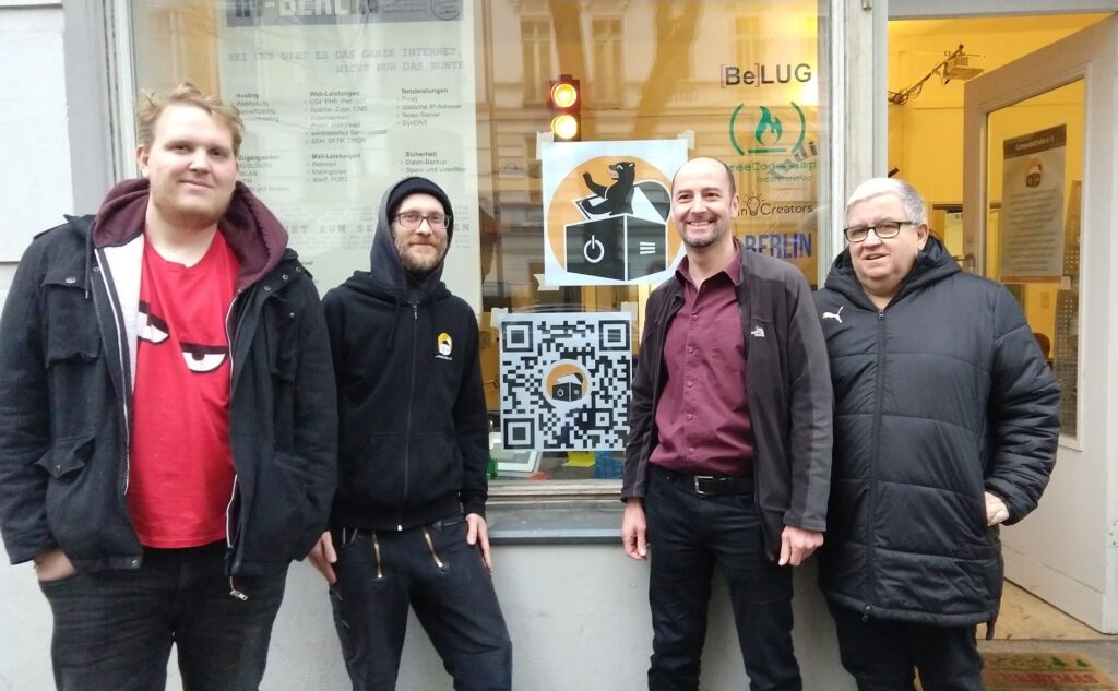 Vier Männer stehen vor dem Schaufenster des geöffneten „IN-Berlin e. V.“-Vereinsraums und lächeln in die Kamera. Am Schaufenster sind auf einem quadratischen Poster das Logo des Berliner Standorts (vor einem runden, orangefarbenen Hintergrund steht der Berliner Bär in einer geöffneten Truhe, die mit dem Symbol für An/Aus und Lüftungsschlitzen bedruckt ist) und auf einem weiteren quadratischen Poster ein QR-Code mit Link zur Homepage der „Computertruhe“ aufgedruckt. Außerdem ein großes Poster vom „IN-Berlin e. V.“ mit der Aufschrift: „Bei uns gibt es das ganze Internet, nicht nur das bunte.“ Einige Schriftzüge weiterer Organisationen, die den Vereinsraum nutzen, sind am Schaufenster erkennbar: „BeLUG, „freeCodeCamp“und „berlinCreators“. Durch die Schaufensterscheibe ist im Raum eine Signalampel zu sehen, die im Moment des Fotos die Farben Rot und Gelb anzeigt. An der geöffneten Eingangstür des Gebäudes ist ein weiteres Poster der „Computertruhe“ erkennbar.