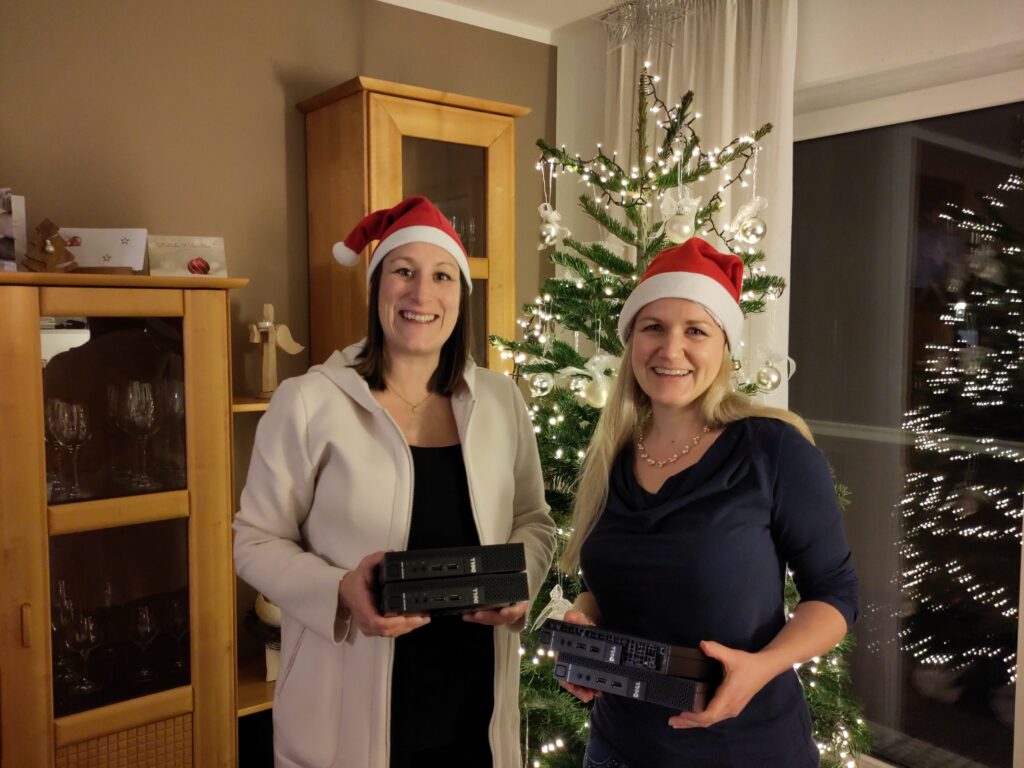 Foto zweier Frauen, die sich in einem weihnachtlich geschmückten Wohnzimmer mit einem Weihnachtsbaum im Hintergrund befinden. Beide tragen rote Zipfelmützen, halten je zwei Mini-Desktops in den Händen und lächeln in die Kamera.