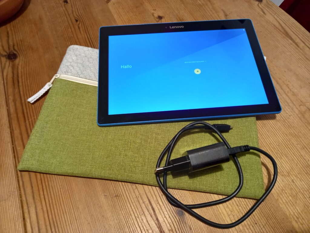 Foto des Tablets in angeschaltetem Zustand. Der Bildschirm zeigt den Einrichtungsassistenten. Eine Hülle und ein Ladegerät liegen dabei.