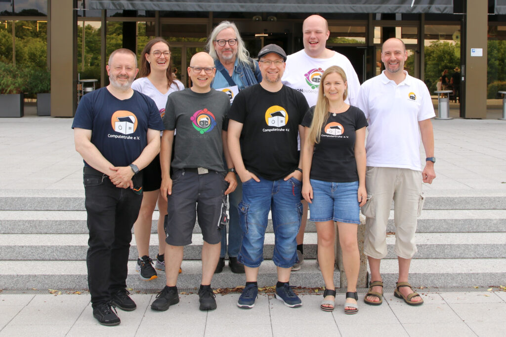 Foto von acht von neun Vorstandsmitgliedern des Vereins „Computertruhe e. V.“. Alle Personen tragen Kleidung, auf der das Vereinslogo abgebildet ist und lächeln in die Kamera.