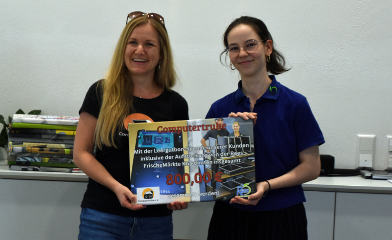 Foto zweier lächelnder Frauen, die gemeinsam eine Leinwand in Händen halten. Darauf sind eine SSD und ein Wagen mit Monitoren abgebildet, hinter dem zwei Menschen stehen. Außerdem steht darauf folgender Text: „Computertruhe – Mit der Leergutbon-Spende unserer Kunden inklusive der Aufstockung von der Rees FrischeMärkte KG konnten insgesamt 800,00 € gesammelt werden!“