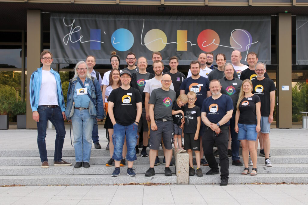 Gruppenfoto, auf welchem 22 Teilnehmer*innen der „CONputertruhe“ sowie zwei Kinder von Vereinsmitgliedern abgebildet sind. Die Personengruppe steht lächelnd auf den Stufen vor dem „Heinz Nixdorf MuseumsForum“ in Paderborn. Die meisten Personen tragen T-Shirts, auf denen das Vereinslogo zu sehen ist.