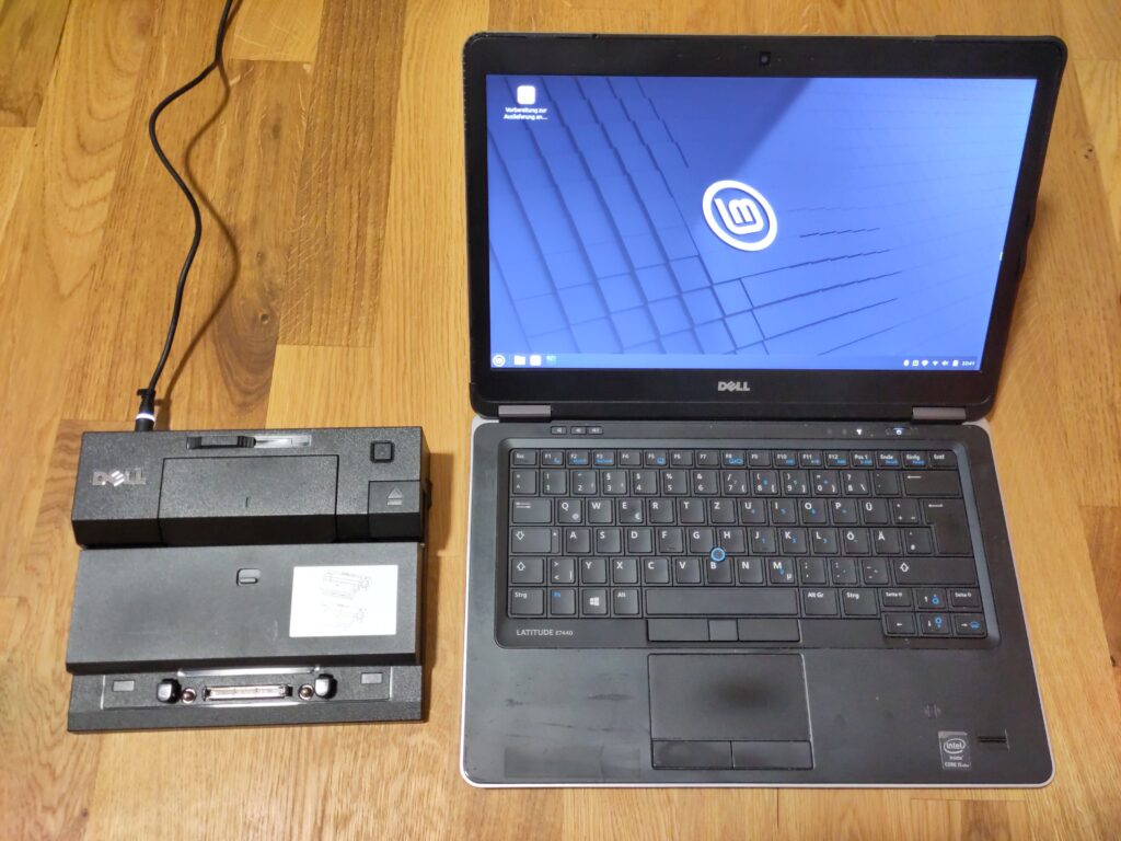 Foto des aufgeklappten Laptops, das den Linux Mint-Desktop anzeigt. Daneben liegen das Dock, das am Netzteil angeschlossen ist. Man kann ein, zwei Schäden am Gehäuse erkennen.