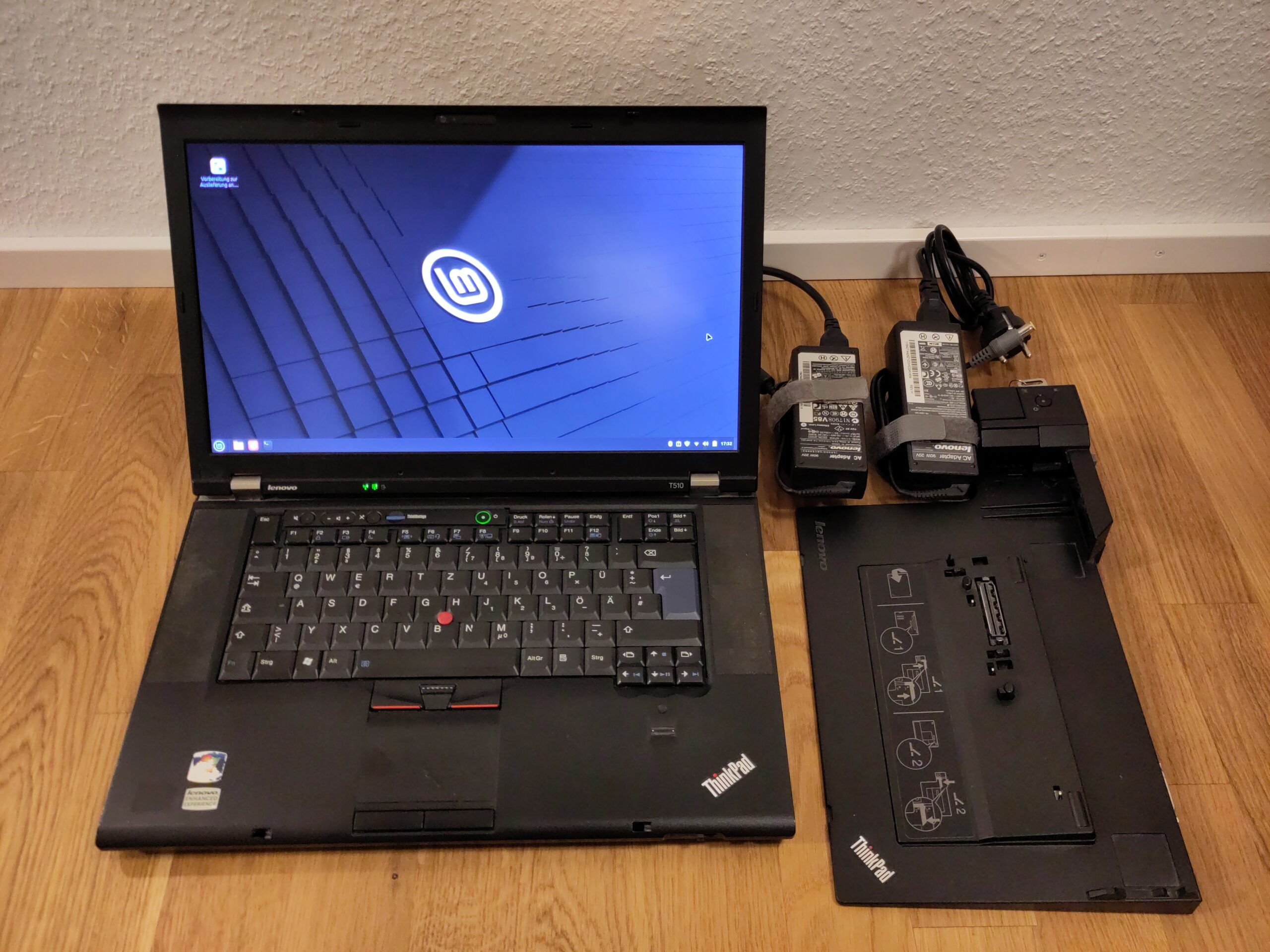 Foto des laufenden Laptops mit üblichen Gebrauchsspuren, das den Linux Mint-Desktop anzeigt. Daneben liegen ein Dock und 2 Netzteile.