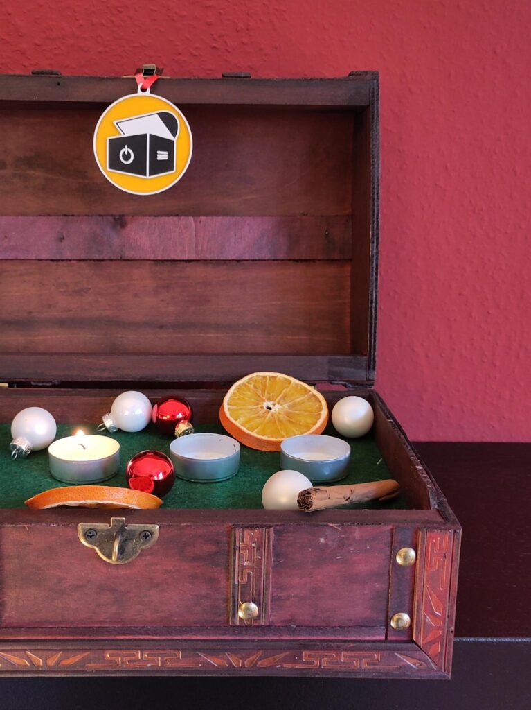 Foto einer aufgeklappten Holztruhe. Darin befinden sich nebeneinander aufgereiht drei Teelichter. Das erste ganz links brennt, während die Lichter in der Mitte und rechts abgebrannt sind. Außerdem ist die Truhe noch geschmückt mit einem Anhänger, der unser Logo zeigt, und kleinen Christbaumkugeln, Orangenscheiben und einer Zimtstange.