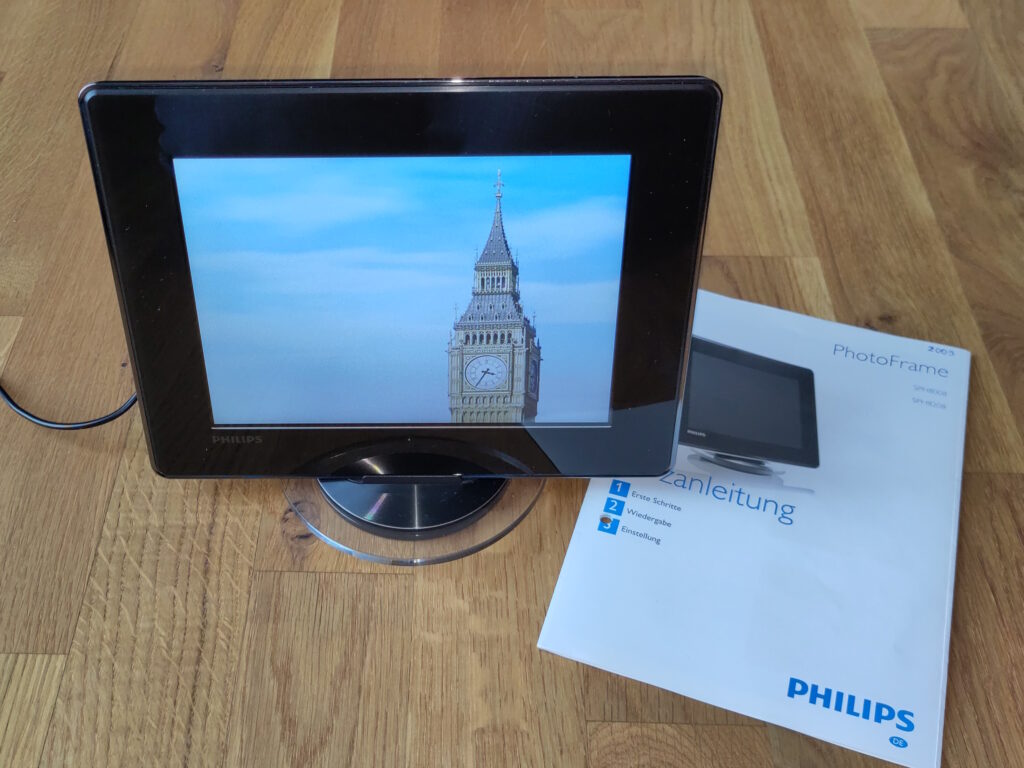 Foto des Bilderrahmens in Betrieb auf einem Ständer inkl. Stromkabel. Daneben liegt die Anleitung.