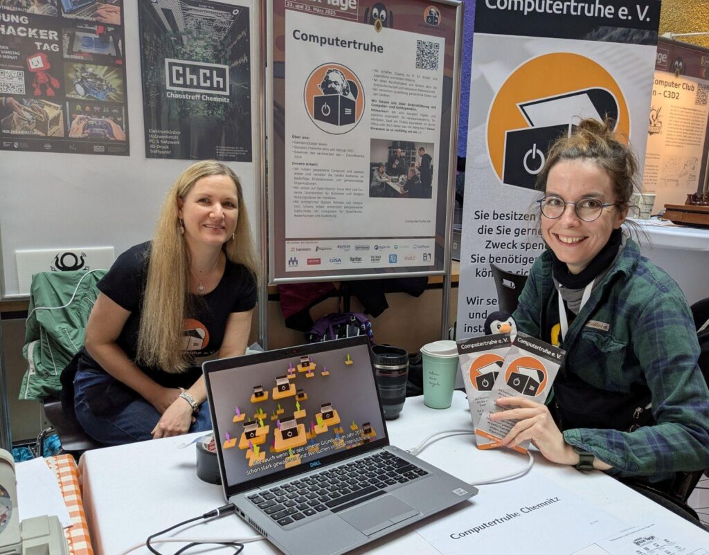 Foto zweier lächelnder Frauen mit Vereins-T-Shirts, die an einem Tisch sitzen. Auf diesem befindet sich zentral ein Laptop, auf dem das Erklärvideo des Vereins abgespielt wird. Die Frau rechts im Bild hält ein paar „Computertruhe“-Faltblätter in der Hand. Im Hintergrund stehen Stellwände mit Postern vom „Chaostreff Chemnitz“ und der „Computertruhe“ und ebenso ein Roll-Up der „Computertruhe“.
