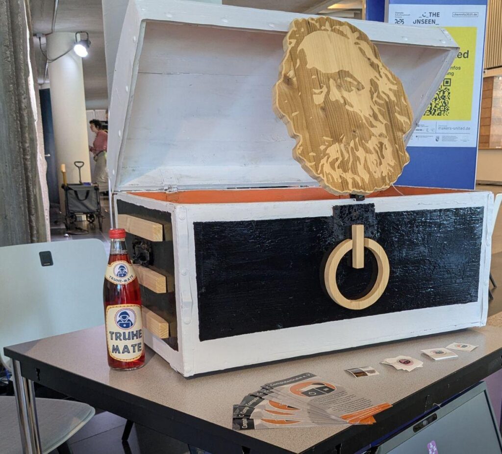 Foto eines Tisches, auf dem sich neben einer „Truhe-Mate“-Flasche und Informations- und Werbematerial des Vereins eine große Holztruhe befindet. Sie ist dem Chemnitzer „Computertruhe“-Standortlogo nachempfunden und darum außen schwarz und weiß bemalt, während das Innere orangefarben ist. Zudem befindet sich vorne das obligatorische Power-Symbol, während an der Seite die Lüftungsschlitze zu sehen sind und ein Holz-Nischel aus der Truhe ragt.
