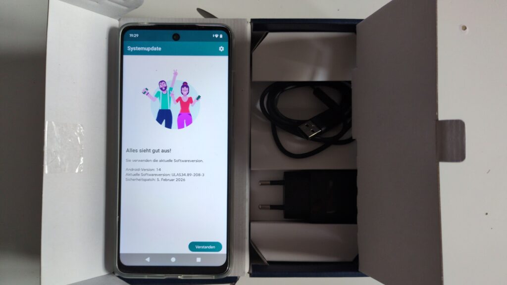 Zu sehen ist der geöffnete Originalkarton des Smartphones. In ihm befindet sich jeweils ein schwarzes USB-Netzteil und ein USB-Ladekabel, sowie das eingeschaltete Smartphone, das in einer durchsichtigen Schutzhülle steckt. Auf dem Smartphone-Bildschirm ist der Menüpunkt "Systemupdate" zu sehen. Zu lesen ist der Text: Alles sieht gut aus! Sie verwenden die aktuelle Softwareversion. Android-Version: 14. Aktuelle Softwareversion: ULAS34.89-208-3. Sicherheitspatch: 5. Februar 2026.