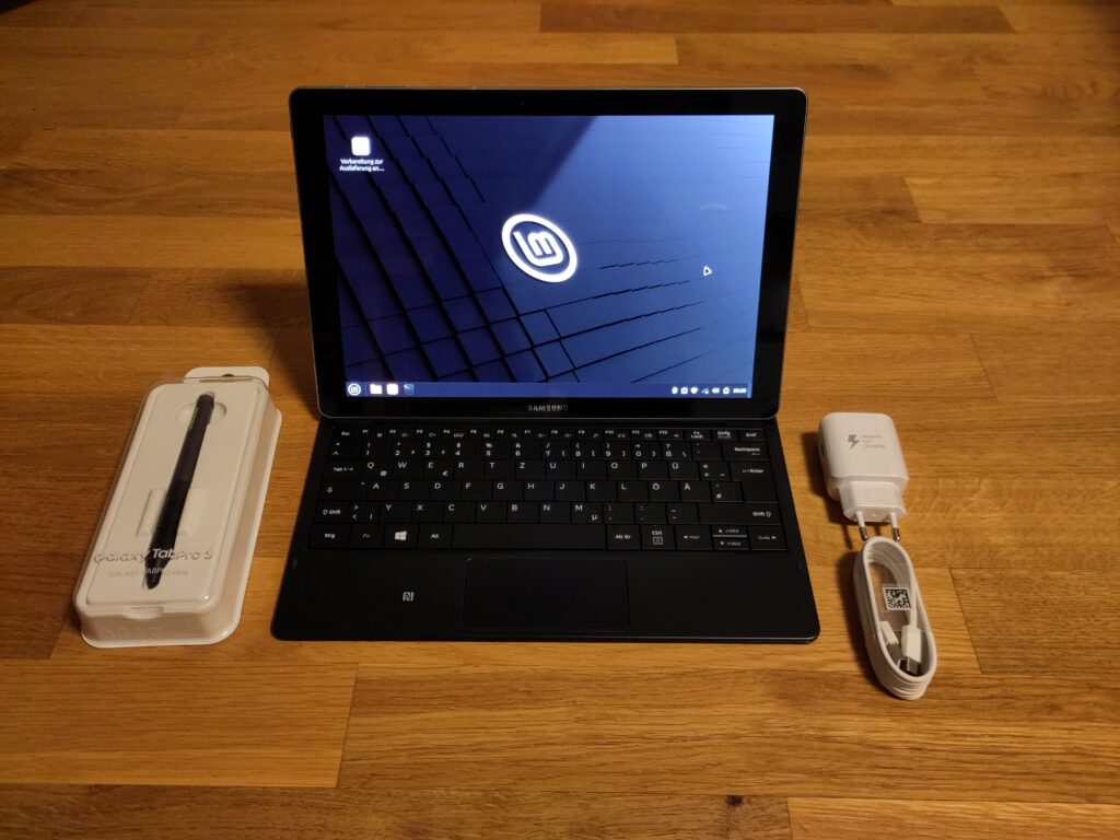 Foto des Gerätes mit laufendem Linux Mint und angesteckt am Tastaturdock. Daneben liegen ein Galaxy TabPro Pen in der Originalverpackung und ein Netzteil nebst USB-Ladekabel. Alles sieht wie neu aus bzw. ist es teilweise auch.