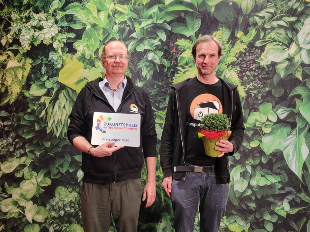 Das Foto zeigt zwei lächelnde Männer vor einer grünen Pflanzentapete, die beide „Computertruhe“-Oberbekleidung tragen. Bei der Person links handelt es sich um Tim Alder, der ein Schild mit der Aufschrift „Zukunftspreis der Sparkasse Chemnitz – Preisträger 2026“ in der rechten Hand vor seinem Körper hält. Die Person rechts ist Robert Köpferl. Er hält in seiner linken Hand einen Blumentopf mit einer grünen Pflanze darin.