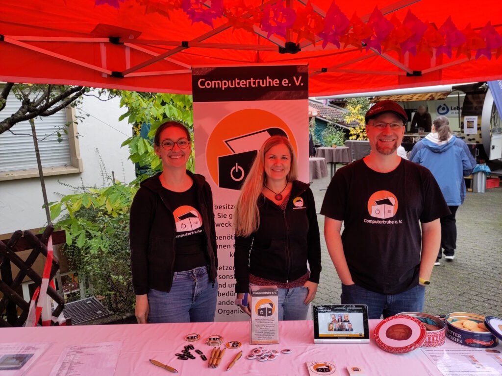 Foto zweier Frauen und eines Mannes unter einem orangefarbenen Pavillon. Die Personen lächeln und tragen schwarze Oberbekleidung, die das Vereinslogo ziert. Vor ihnen befindet sich ein langer Tisch mit diversem Info- und Werbematerial, einem Tablet, das eine Präsentation zeigt, und mit zwei geöffneten Keksdosen. Hinter ihnen steht ein Roll-up.