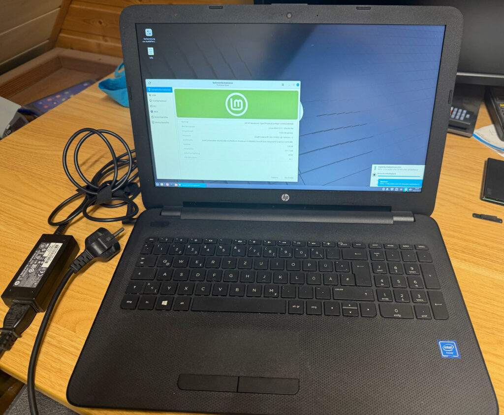 15,6" schwarzer Laptop, der den Linux Mint-Desktop anzeigt. Es liegt das Netzteil daneben, welches aber nicht an einer Steckdose angeschlossen ist.