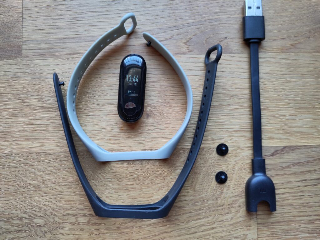 Foto des Fitnesstrackers, einem grauen und schwarzen Armband, 2 Ersatzarmbandknöpfchen und dem Ladekabel.
