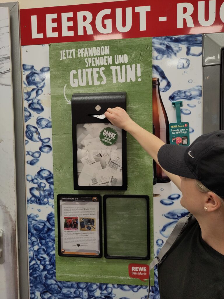 Foto, das eine Wand neben einem Pfandrückgabeautomaten zeigt, an dem eine abschließbare Box mit transparenter Front hängt. Darin befinden sich jede Menge Pfandbons. Über der Box steht an der Wand der Satz „Jetzt Pfandbon spenden und Gutes tun!“. Eine Frau steckt im Moment der Aufnahme einen Bon in den Einwurfschlitz der Box. Unter der Box hängt außerdem ein DIN A4-großes Plakat, welches die „Computertruhe“ vorstellt.
