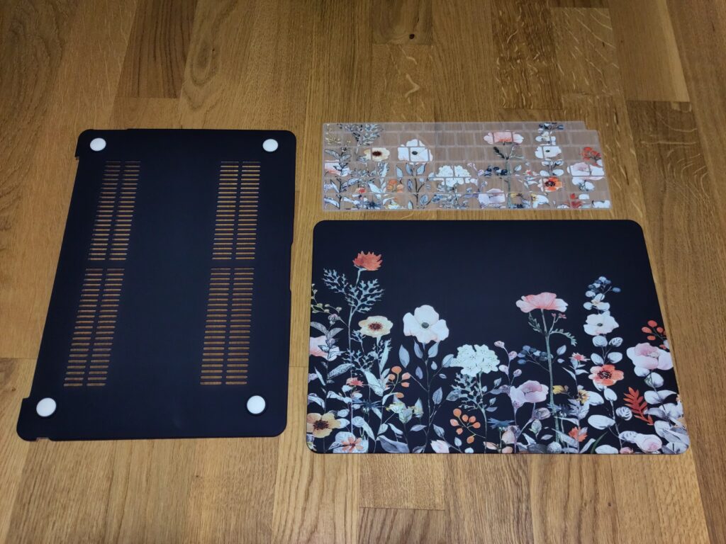 Foto der dunkelblauen Hülle bestehend aus der Unterseite und der Oberseite. Letztere ist mir bunten Blumen verziert. Ebenfalls sichtbar ist eine Tastaturmatte aus Silikon, die mit demselben Blumenmuster verziert ist.
